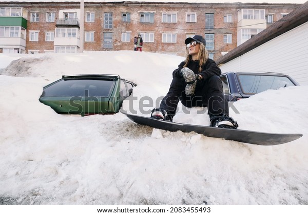 Portrait of snowboarder sitting in\
city near cars under deep snow. Urban street\
snowboarding