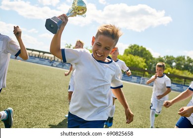 Portrait Of Soccer Team Captain Running Across Field Holding Golden Cup, With His Teammates Cheering In Background