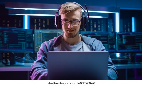 Portrait of Software Developer / Hacker / Gamer Wearing Glasses and Headset Sitting at His Desk and Working / Playing on Laptop. In the Background Dark High Tech Environment with Multiple Displays.