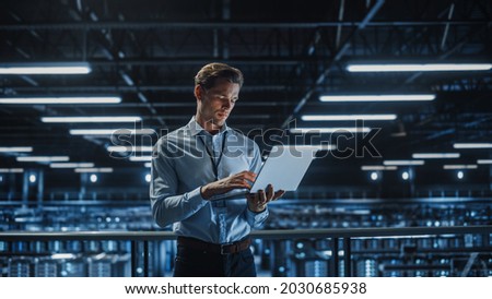Portrait of IT Specialist Uses Laptop in Data Center. Server Farm Cloud Computing Facility with Male Maintenance Administrator Working. Cyber Security and Network Protection.