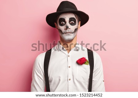 Portrait of spooky handsome guy made makeup for Halloween event, has image of vampire or ghost, red rose flower in pocket of white shirt, wears black hat, has scary look, dressed in zombie attire