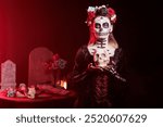 Portrait of spooky lady holding creepy skull during Dia de los Muertes indigenous rites of honoring the dead. Woman wearing flower crown participating in Day of the Dead event in honor of the deceased