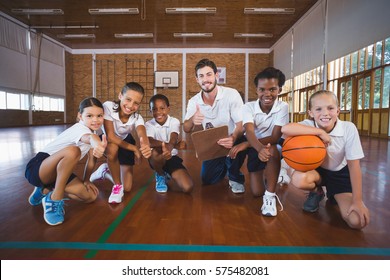 Portrait Of Sports Teacher And School Kids Showing Thumbs Up In Basketball Court At School Gym