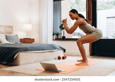 Portrait of sporty African American woman having online training at bedroom and squatting, using laptop at home. Wellness and healthy lifestyle concept - Powered by Shutterstock