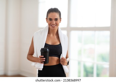 Portrait of sporty beautiful smiling lady in sportswear and white towel on neck holding shaker with healthy drink, whey protein, fresh water or coffee and posing looking at camera, standing by window - Powered by Shutterstock