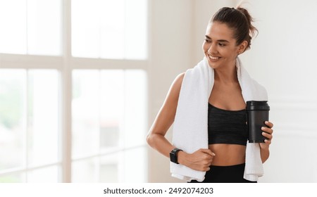 Portrait of sporty beautiful smiling lady in sportswear and white towel on neck holding shaker with healthy drink, whey protein, fresh water or coffee and looking away at free copy space banner mockup - Powered by Shutterstock