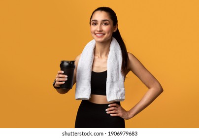Portrait of sporty beautiful smiling woman in black sportswear and white towel on neck holding shaker with healthy drink, whey protein, fresh water or coffee isolated over orange studio background