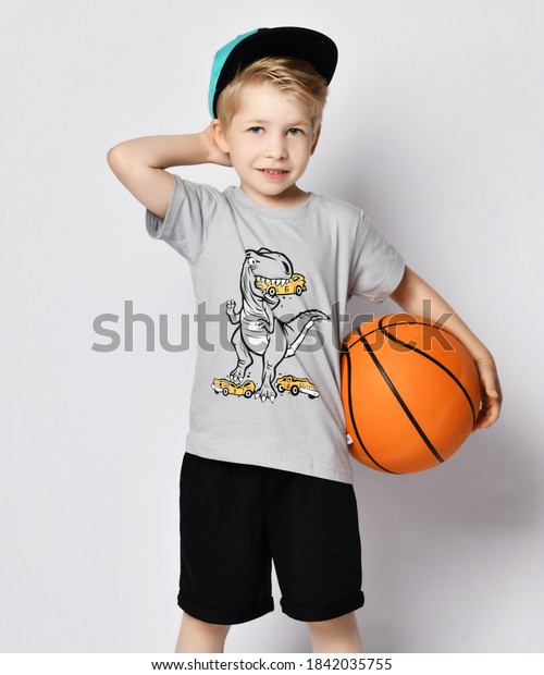 Portrait of sporty blond\
kid boy 6-7 y.o. in cap, blue t-shirt with dinosaur eating cars\
print and black shorts standing with basketball ball holding hand\
at his nape head