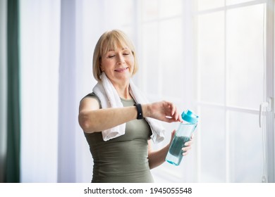 Portrait of sporty senior woman with bottle of water checking her watch or fitness tracker indoors. Athletic mature lady staying hydrated during sports training, leading healthy lifestyle - Powered by Shutterstock