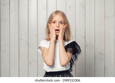 Portrait Of Startled Blonde Teen Girl Raising Hands And Gasping Worried, Staring Shocked And Confused At Camera. Studio Shot, Light Wooden Background