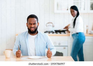 Portrait Of Starving Angry Black Man Waiting For Food At Table In Kitchen, Hungry African American Husband Sitting With Empty Plate And Looking At Camera, His Wife Cooking Lunch On Background