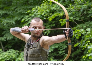 Portrait Of A Stern Man Shooting An Arrow From A Bow In The Forest. Concept: Men's Hobbies, Survival In Difficult Conditions, Hunting And Recreation.