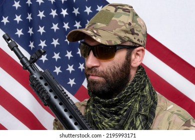 Portrait Of A Stern Military Man 30-35 Years Old With A Rifle On His Shoulder, Wearing Dark Glasses Against The Background Of The American Flag.Concept: Military Action, Soldier Of Fortune, US Army.