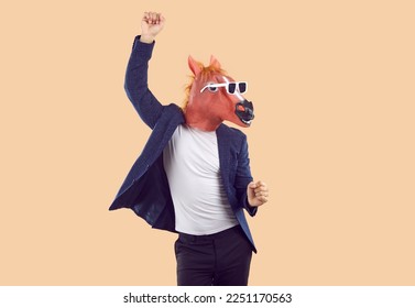 Portrait of strange man wearing T shirt, suit jacket, funny horse mask and sunglasses having fun and dancing isolated on beige background. Fun, party, humor concept