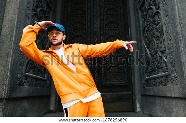 Portrait of a street dancer standing against the\
background of an old building and dancing hip hop, wearing a bright\
sports suit. Young man shows hip hop performance on the street.\
Street dancing.