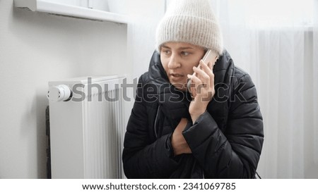 Portrait of stressed brunette woman in winter coat and woolen hat calling service to repair broken heater radiator.