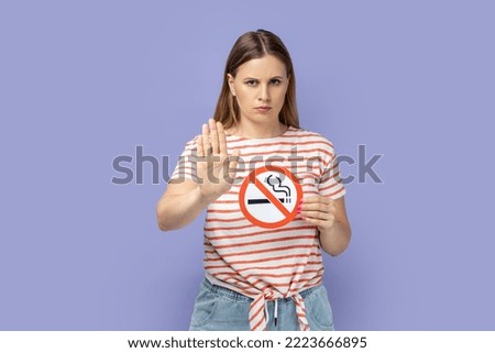 Portrait of strict attractive blond woman wearing striped T-shirt holding no smoking sign and showing stop gesture with her palm. Indoor studio shot isolated on purple background.