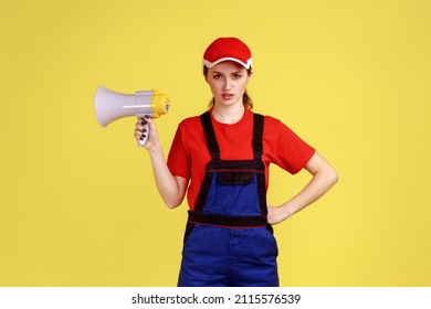 Portrait Of Strict Bossy Worker Woman Holding Megaphone In Hands, Keeps Hand On Hip, Looking At Camera With Serious Expression, Wearing Overalls. Indoor Studio Shot Isolated On Yellow Background.