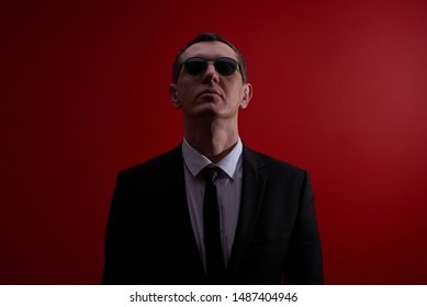 Portrait Of A Strict Serious Adult Man In A Suit And Black Glasses On A Red Studio Background. Face Control, Night Club Security