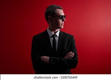 Portrait Of Strict Serious Adult Man In Suit And Black Glasses Looks Away With Crossed Arms On Red Studio Background. Face Control, Night Club Security