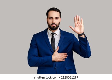 Portrait Of Strict Serious Bearded Man Raising One Arm And Putting On Chest Another Making Oath, Swearing, Wearing Official Style Suit. Indoor Studio Shot Isolated On Gray Background.