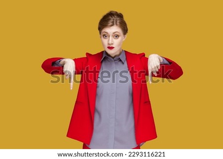 Portrait of strict serious woman with red lips standing pointing both index fingers down, saying here and right now, wearing red jacket. Indoor studio shot isolated on yellow background.