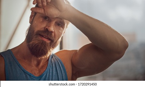 Portrait Of Strong Bearded Male Athlete Wearing Sleeveless Shirt Wipes Sweat From His Forehead With His Muscular Hand. Handsome Man After Hardcore Exercise And Training. Man Gets Job Done