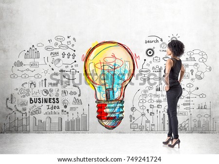 Portrait of a strong and independent African American woman wearing black and standing with her hands on the waist. Concrete wall with a business plan sketch and a large light bulb