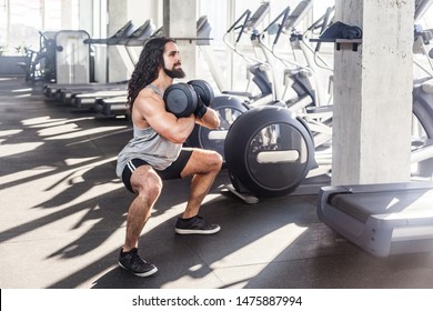 Portrait Of Strong Young Adult Man With Long Curly Hair Handsome Athlete Working Out In Gym, Static Squat And Holding Two Dumbbells, Doing Exercises For Legs And Squatting. Indoor, Looking Away