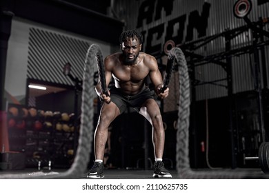 Portrait of strong young african man exercising with battle ropes during workout in modern gym, alone. indoors. active male having intense cross fit training, in dark sports club. fitness concept - Shutterstock ID 2110625735