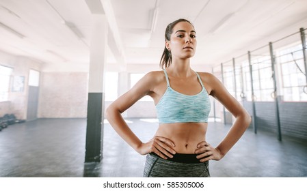 Portrait Of Strong Young Woman Standing In Gym With Her Hands On Hips. Caucasian Female Fitness Model Looking At Camera.