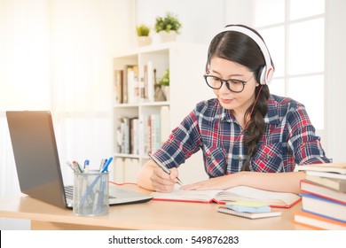 Portrait of a student learning on line with headphones and laptop taking notes in a notebook sitting at her desk at home. mixed race asian chinese model.