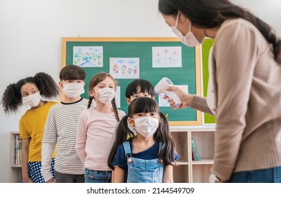 Portrait of students in medical mask in classroom, covid-19 outbreak. Teacher kids with thermometer at preschool entrance. Social distancing, coronavirus prevention. Temperature check in at school.  - Shutterstock ID 2144549709