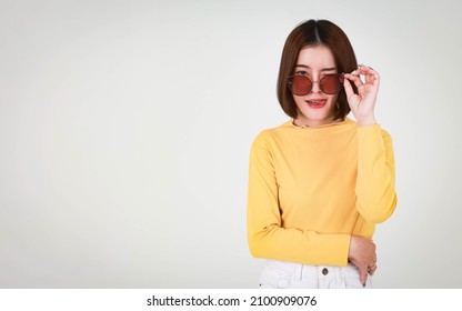 Portrait Studio Cutout Shot Of Asian Young Beautiful Stylish Short Hair Female Model In Yellow Long Sleeve Shirt Standing Crossed Arm Holding Hand Adjusting Fashion Sunglasses On White Background.