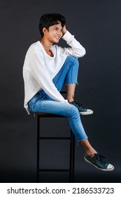 Portrait Studio Full Body Shot Of Asian Young LGBTQ Gay Glamour Handsome Male Model In Casual White Shirt Jeans Sneakers Sitting On Tall Chair Laughing Holding Hand Cover Mouth On Black Background.