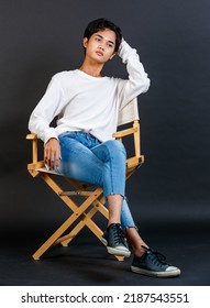 Portrait Studio Full Body Shot Of Asian Young LGBTQ Gay Glamour Handsome Bisexual Homosexual Male Model In Casual Outfit Sitting Crossed Legs On Wooden Chair Posing Gesturing On Black Background.