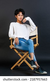 Portrait Studio Full Body Shot Of Asian Young LGBTQ Gay Glamour Handsome Bisexual Homosexual Male Model In Casual Outfit Sitting Crossed Legs On Wooden Chair Posing Gesturing On Black Background.