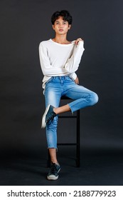 Portrait Studio Full Body Shot Of Asian Young LGBTQ Gay Glamour Handsome Male Model In Casual White Shirt Jeans Sneakers Sitting On Tall Chair Laughing Holding Hand Cover Mouth On Black Background.