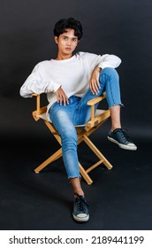 Portrait Studio Full Body Shot Of Asian Young LGBTQ Gay Glamour Handsome Bisexual Homosexual Male Model In Casual Outfit Sitting Crossed Legs On Wooden Chair Posing Gesturing On Black Background.