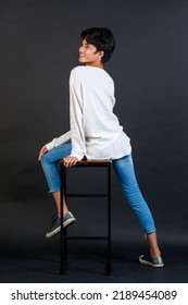 Portrait Studio Full Body Shot Of Asian Young LGBTQ Gay Glamour Handsome Male Model In Casual White Shirt Jeans Sneakers Sitting On Tall Chair Laughing Holding Hand Cover Mouth On Black Background.