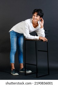 Portrait Studio Full Body Shot Of Asian Young LGBTQ Gay Glamour Handsome Male Model In Casual White Shirt Jeans Sneakers Sitting On Tall Chair Laughing Holding Hand Cover Mouth On Black Background.