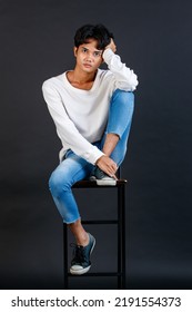 Portrait Studio Full Body Shot Of Asian Young LGBTQ Gay Glamour Handsome Male Model In Casual White Shirt Jeans Sneakers Sitting On Tall Chair Laughing Holding Hand Cover Mouth On Black Background.