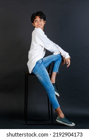 Portrait Studio Full Body Shot Of Asian Young LGBTQ Gay Glamour Handsome Male Model In Casual White Shirt Jeans Sneakers Sitting On Tall Chair Laughing Holding Hand Cover Mouth On Black Background.