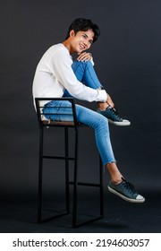 Portrait Studio Full Body Shot Of Asian Young LGBTQ Gay Glamour Handsome Male Model In Casual White Shirt Jeans Sneakers Sitting On Tall Chair Laughing Holding Hand Cover Mouth On Black Background.