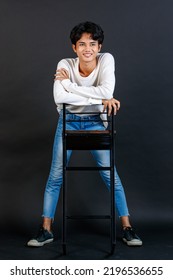 Portrait Studio Full Body Shot Of Asian Young LGBTQ Gay Glamour Handsome Male Model In Casual White Shirt Jeans Sneakers Sitting On Tall Chair Laughing Holding Hand Cover Mouth On Black Background.