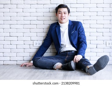 Portrait Studio Shot Asian Fashionable Handsome Successful Professional Male Businessman In Casual Blue Suit Turtle Neck Shirt Sit Stretch Legs On Floor Look At Camera On White Brick Wall Background.