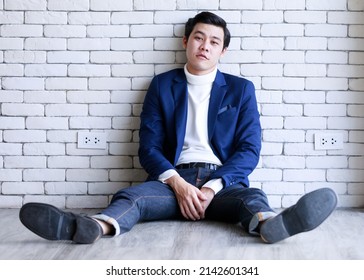 Portrait Studio Shot Asian Fashionable Handsome Successful Professional Male Businessman In Casual Blue Suit Turtle Neck Shirt Sit Stretch Legs On Floor Look At Camera On White Brick Wall Background.