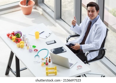 Portrait Studio Shot Asian Male Professional Nutritionist Dietitian Doctor In White Lab Coat And Stethoscope Sitting Holding Pen Smiling At Working Desk Full Of Fruits Vegetable Nutrition Supplements.