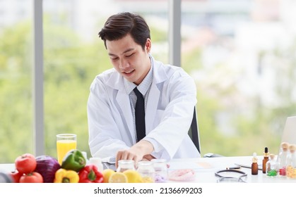 Portrait Studio Shot Asian Male Professional Nutritionist Dietitian Doctor In White Lab Coat And Stethoscope Sitting Holding Pen Smiling At Working Desk Full Of Fruits Vegetable Nutrition Supplements.