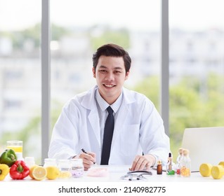 Portrait Studio Shot Asian Male Professional Nutritionist Dietitian Doctor In White Lab Coat And Stethoscope Sitting Holding Pen Smiling At Working Desk Full Of Fruits Vegetable Nutrition Supplements.